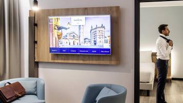 Una camera d'albergo con una TV che mostra una vista sulla città, una poltrona blu, una borsa in pelle marrone e un pavimento in legno.