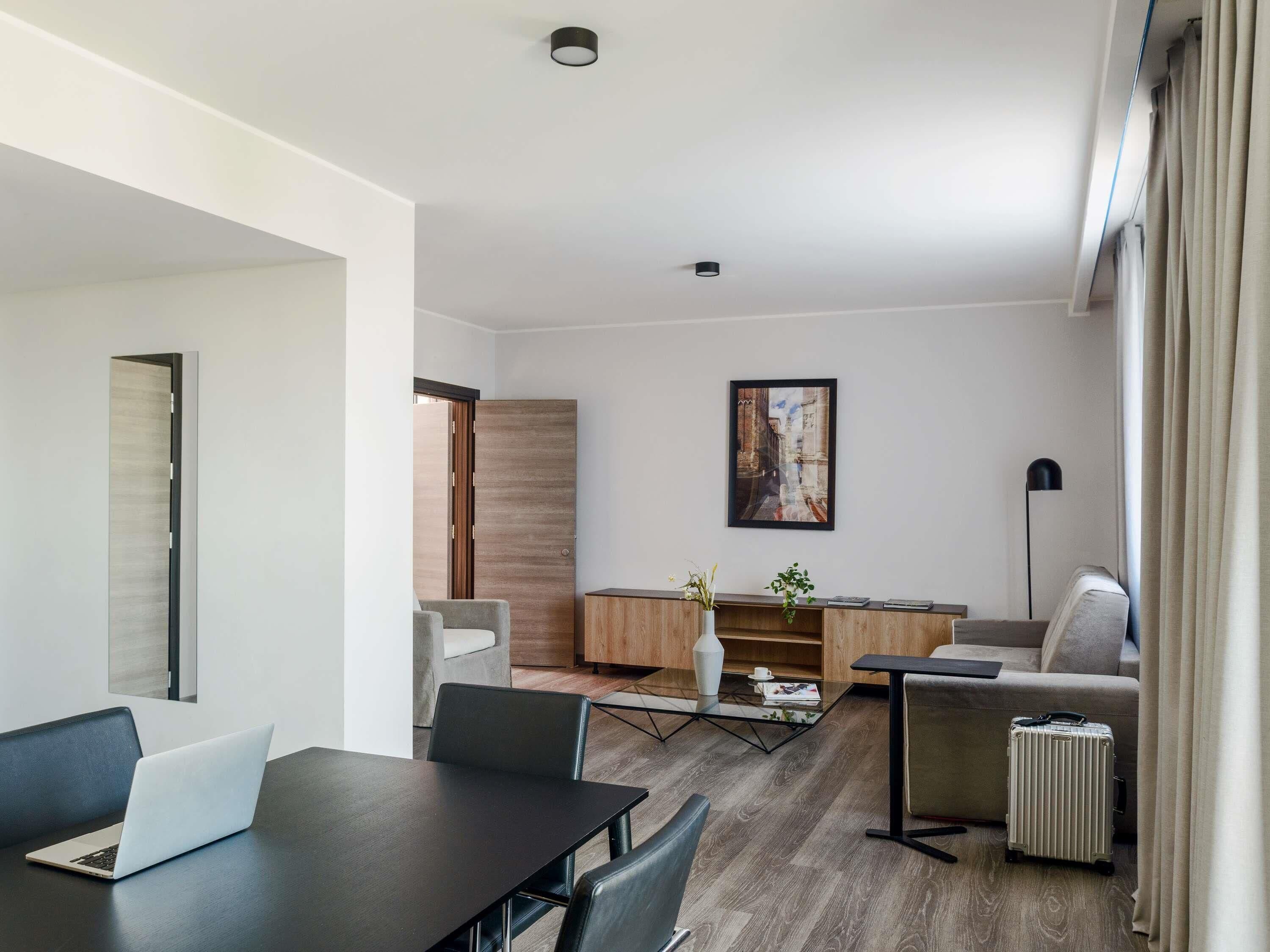 Ein modernes Hotelzimmer mit Essbereich, einer Couch, einem Couchtisch, einer Lampe und einem Koffer.