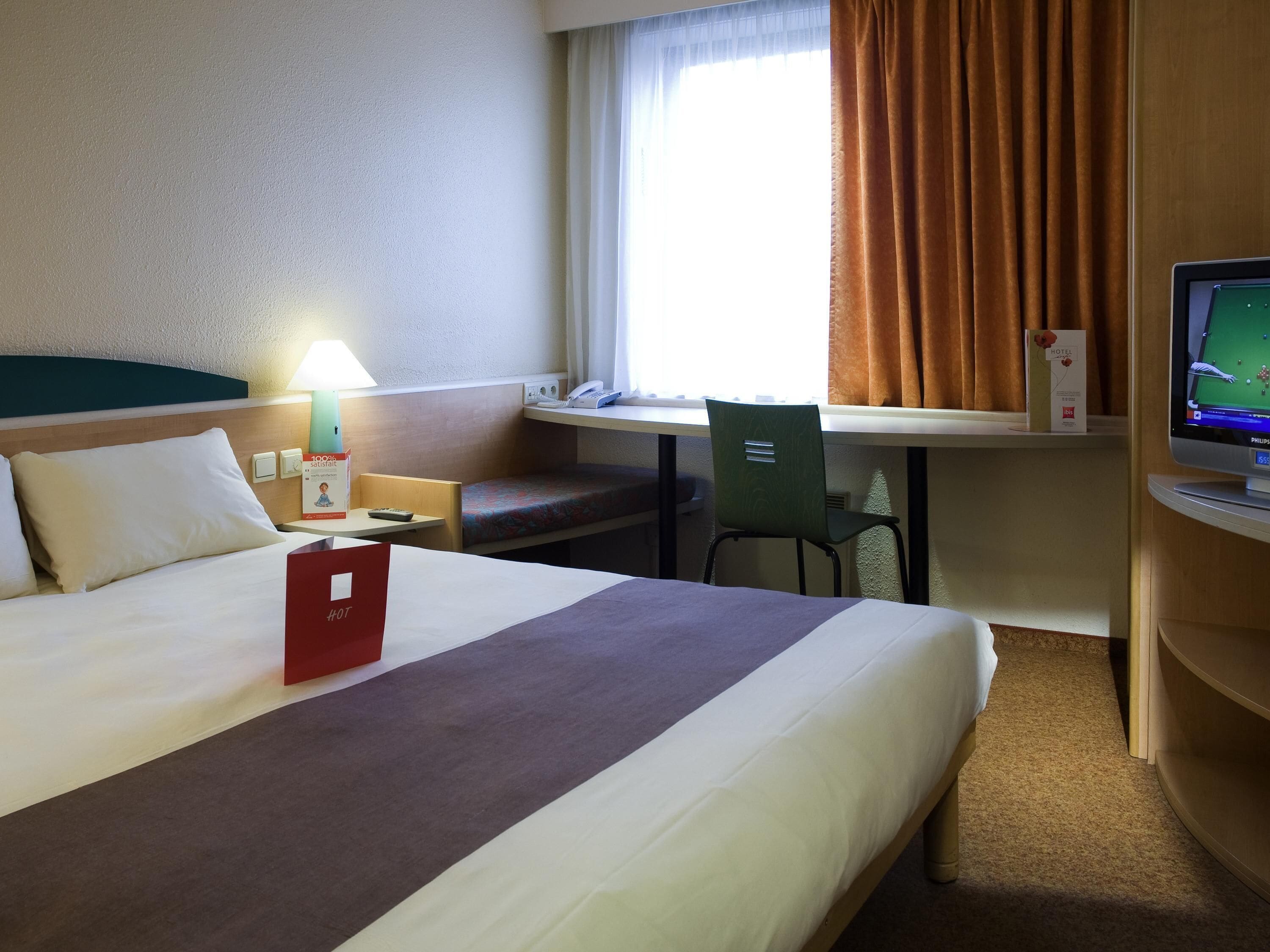 Una habitación de hotel con cama, escritorio, silla, televisor y ventana con cortinas.