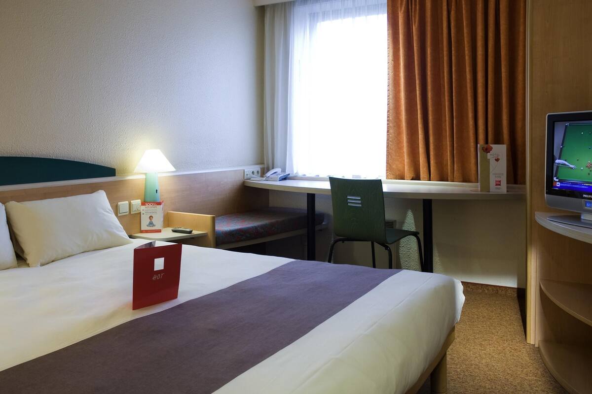 Una camera d'albergo con un letto, una scrivania, una sedia, una televisione e una finestra con tende.