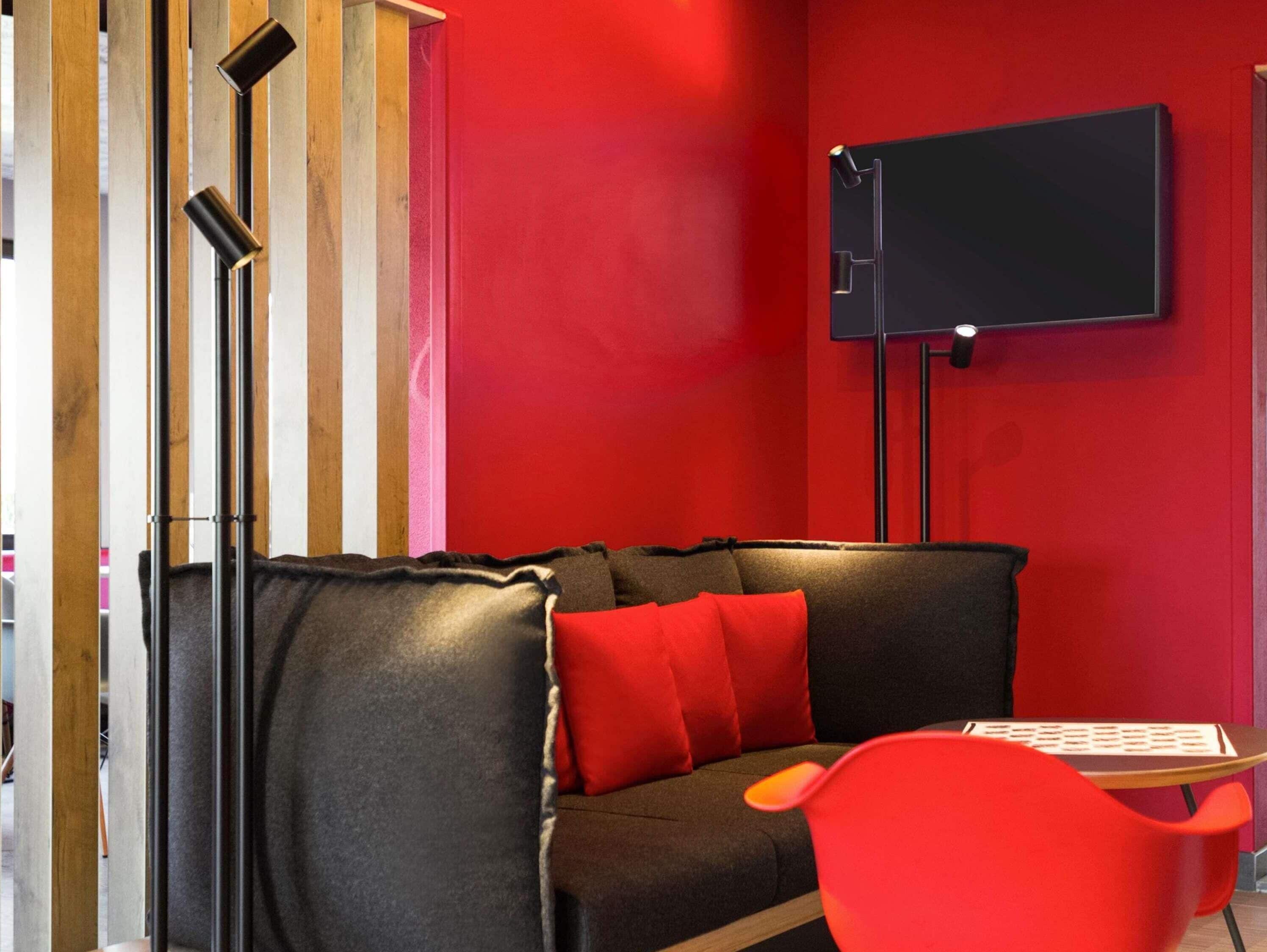 Quarto moderno com parede vermelha, sofá preto com almofadas vermelhas, estante de madeira e televisão de tela plana fixada na parede.