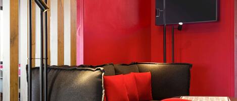 A modern room with a red wall, a black sofa with red cushions, a wooden bookshelf, and a flat-screen TV mounted on the wall.