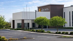 A modern hotel with a flat roof, a sign reading 'La Quinta,' and a parking lot with trees and shrubs.