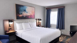 A hotel room with a bed, bedside tables, a desk, a chair, a painting, and a window with curtains.