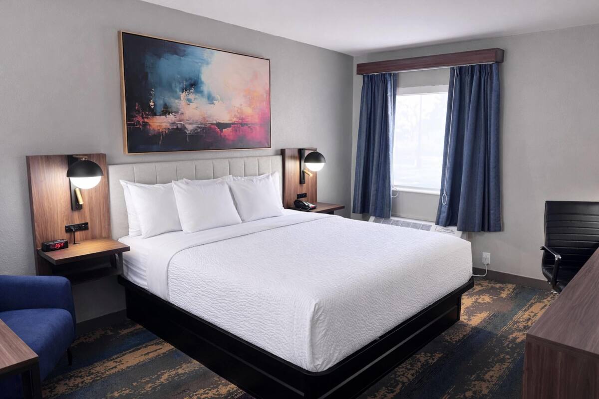A hotel room with a bed, bedside tables, a desk, a chair, a painting, and a window with curtains.