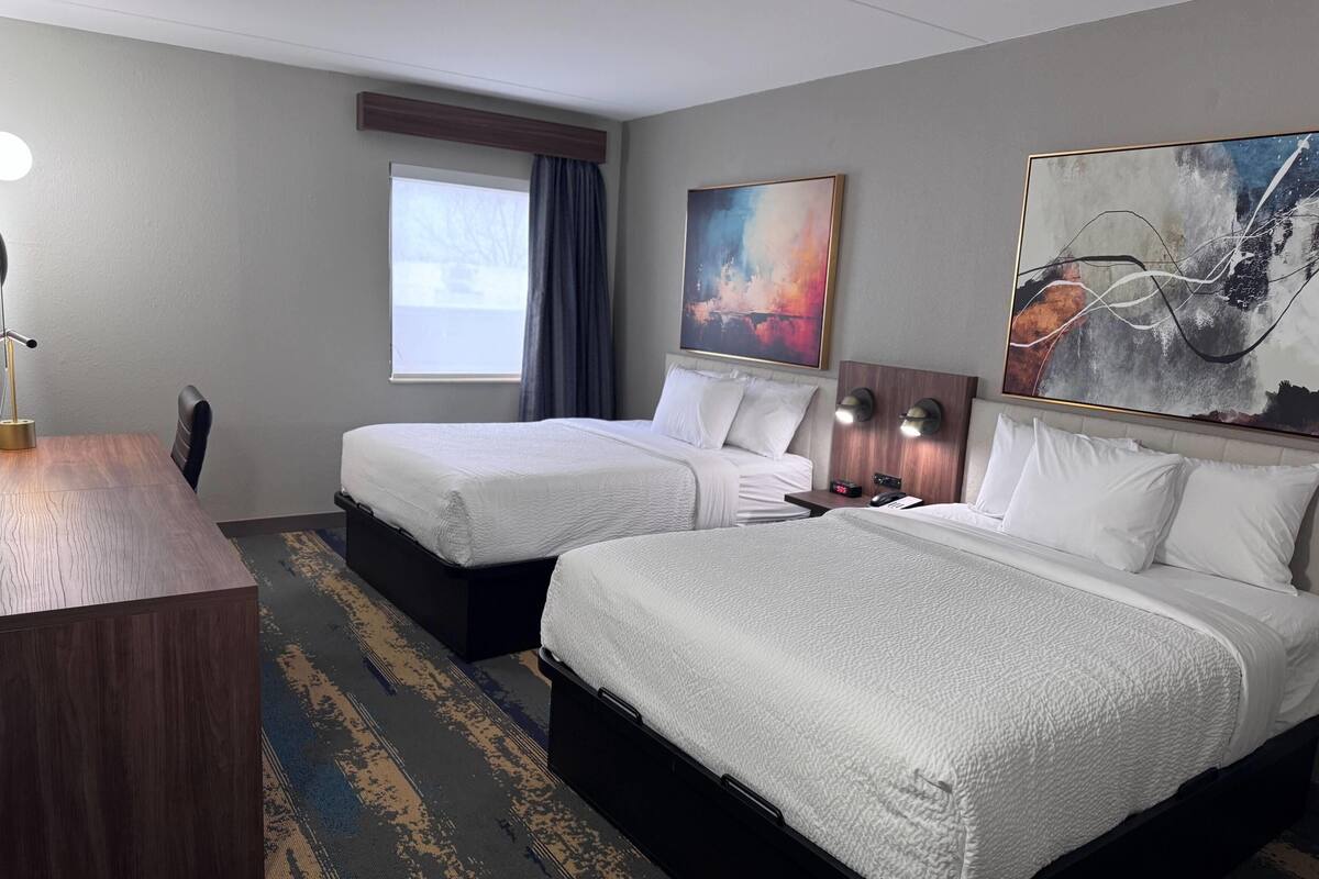 A hotel room with two beds, a desk, a chair, a window with curtains, and artwork on the walls.