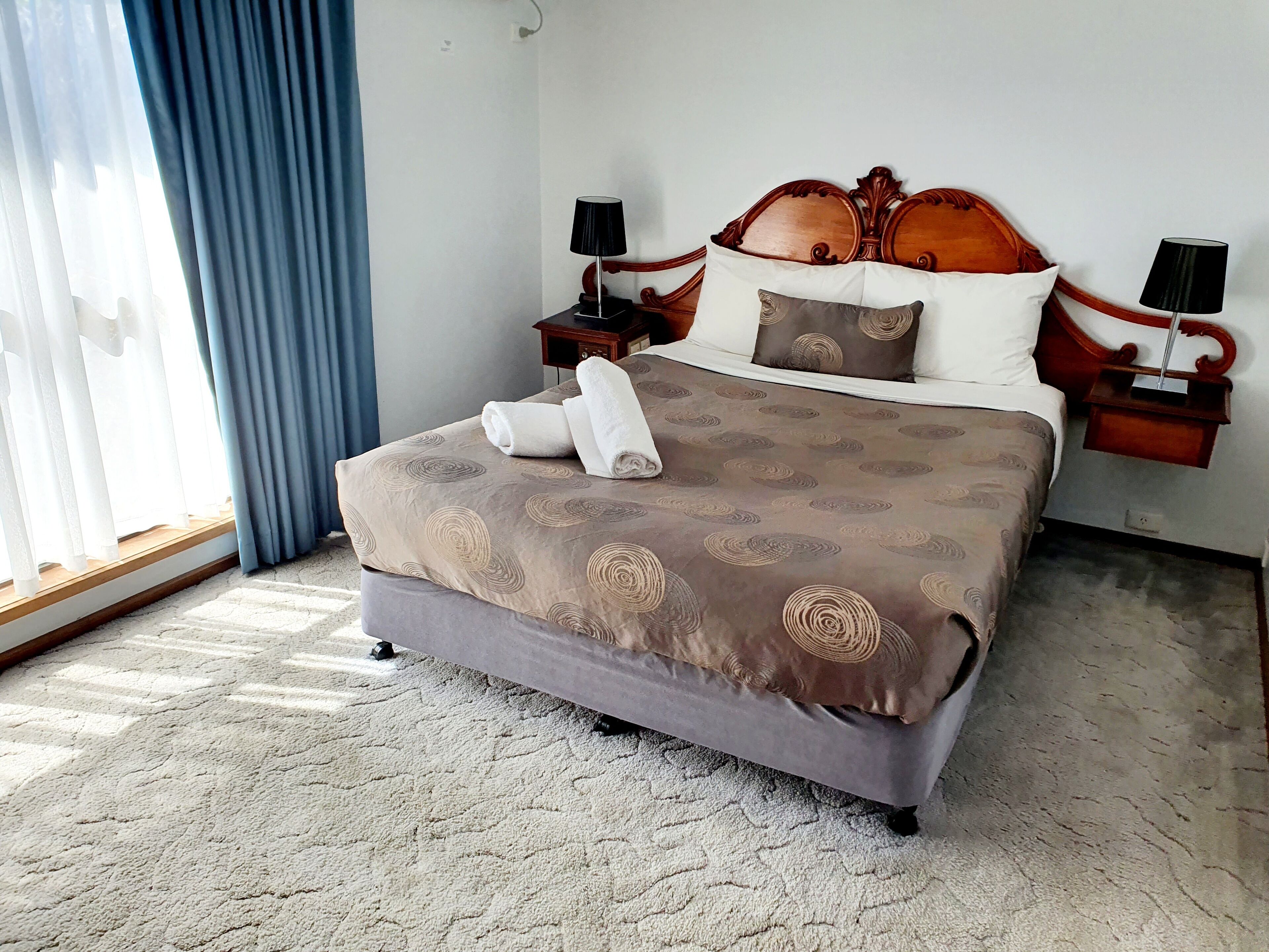 A neatly made bed with a patterned bedspread, two bedside tables with lamps, a window with blue curtains, and a carpeted floor.