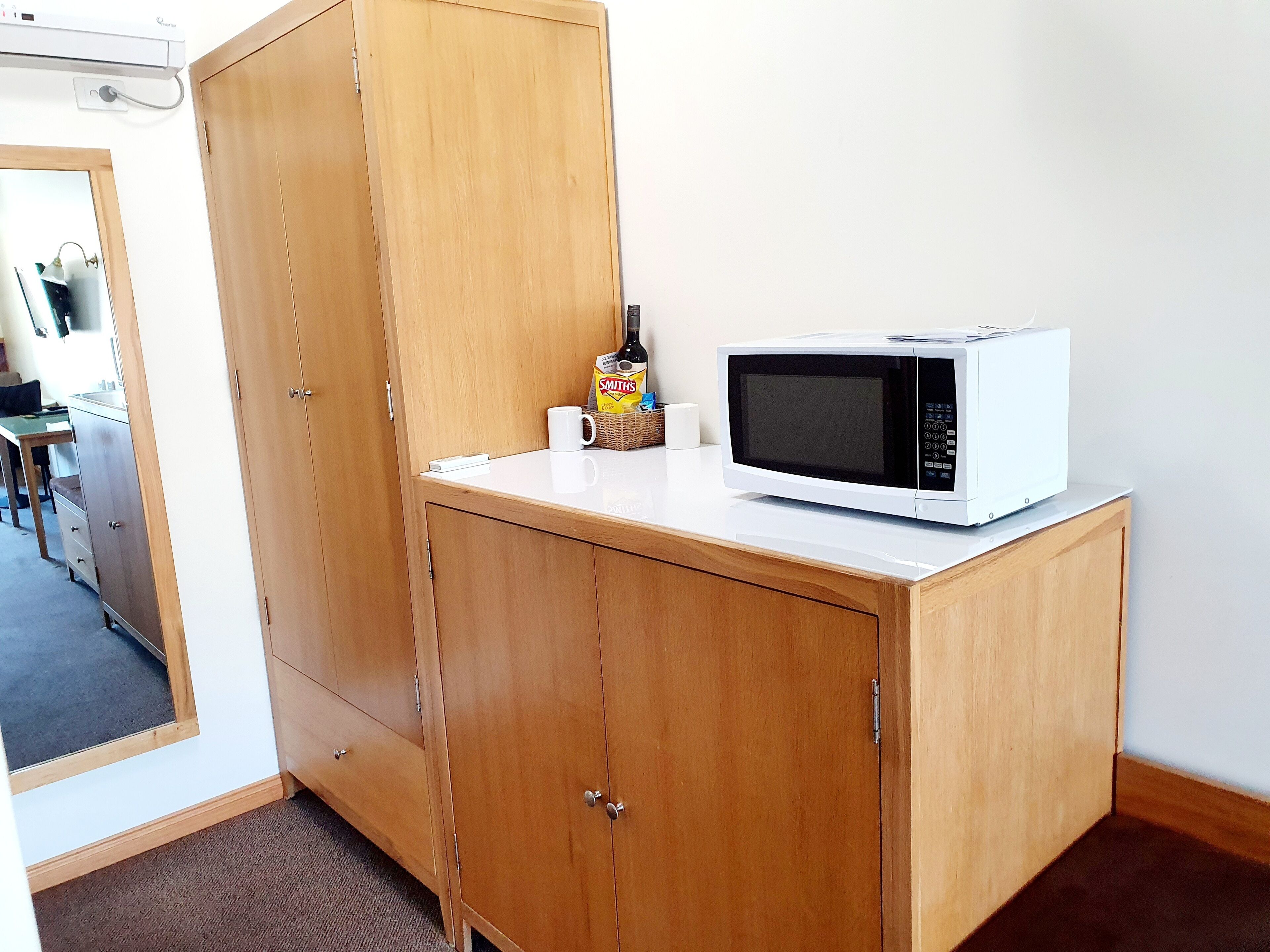 A small kitchenette with a microwave on a white countertop, a wooden cabinet, and a wicker basket with snacks.