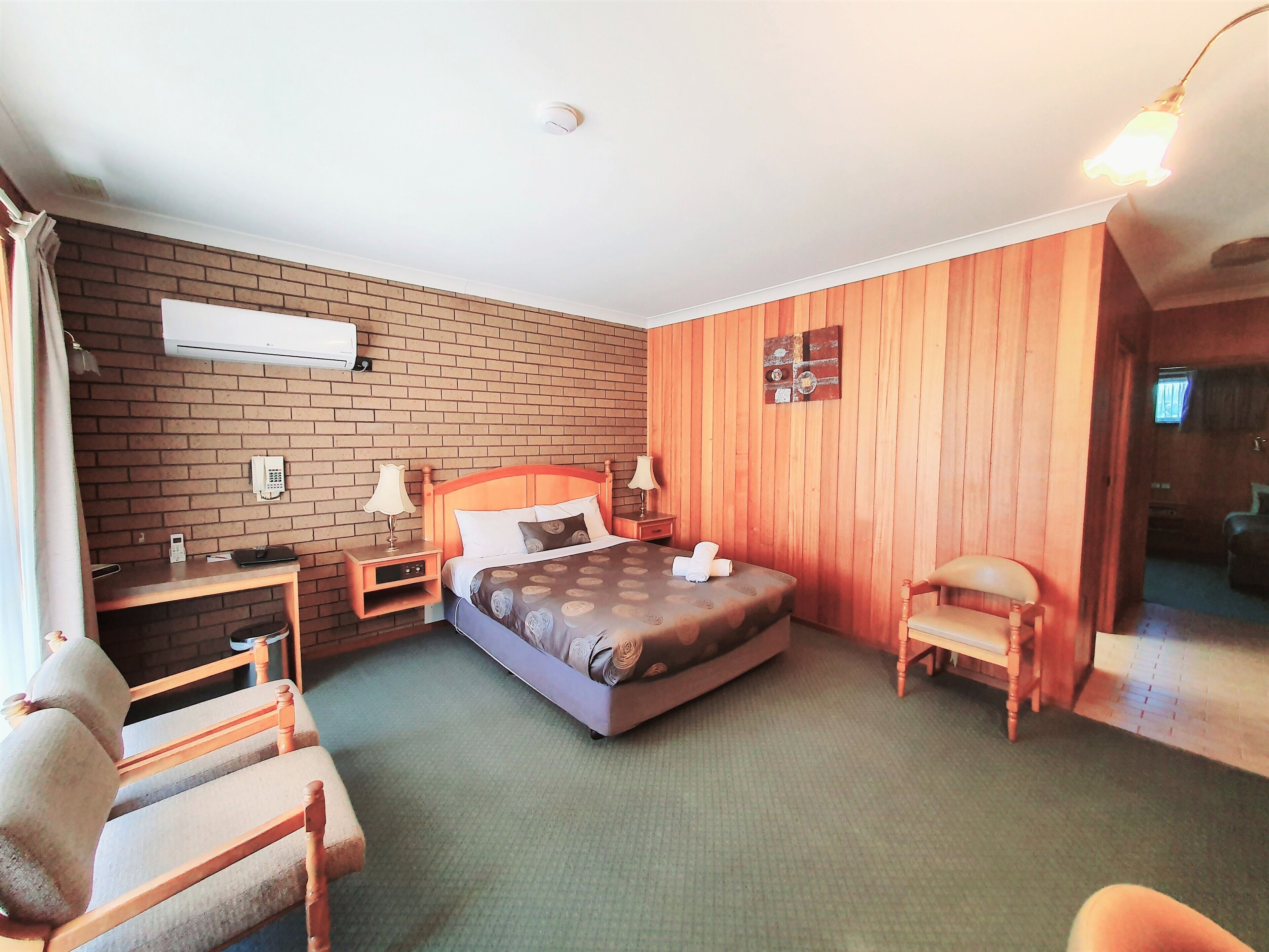 Hotelkamer met een bakstenen muur, houten lambrisering, een bed met dekbed, een bureau met lamp, een stoel en een nachtkastje.