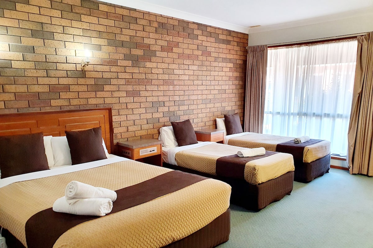 A hotel room with three beds, a brick wall, a wooden headboard, a nightstand, and a window with curtains.