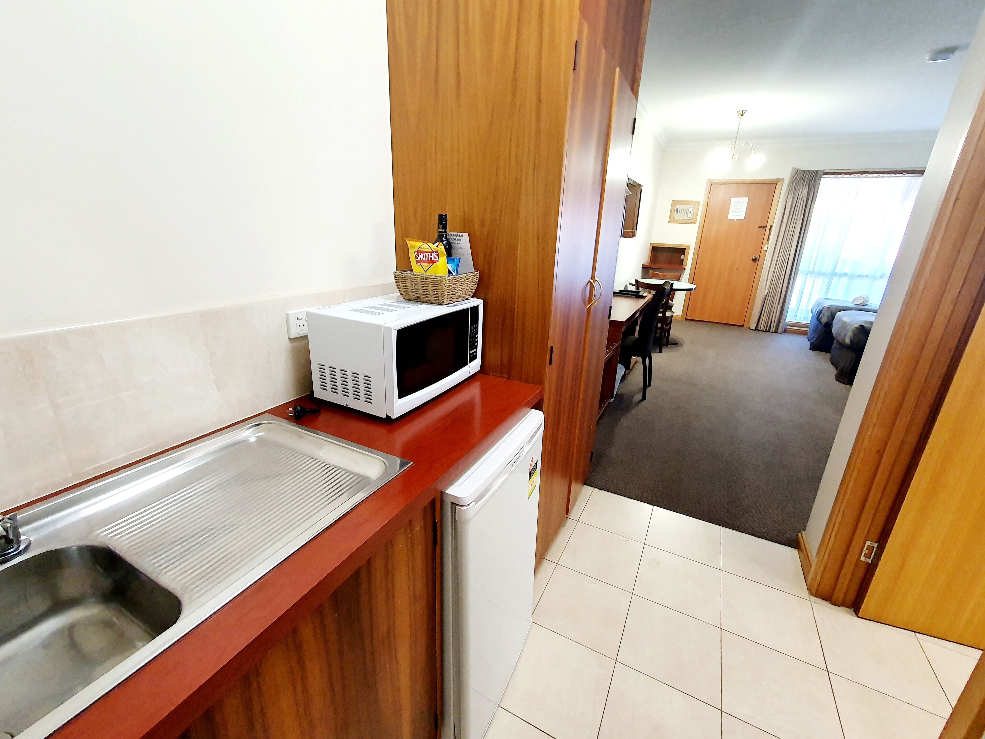A kitchenette with a microwave, a sink, and a small refrigerator. There is a wicker basket with snacks and a bottle on the microwave. The room has a wooden door and a view of a living area with a sofa and a table.