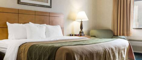 Habitación de hotel con una cama grande, una mesita de noche con lámpara, una silla verde, un cuadro en la pared y una ventana con cortinas.
