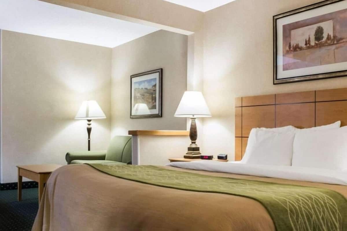 A hotel room with a bed, two bedside lamps, a chair, a small table, a framed picture on the wall, and a headboard.