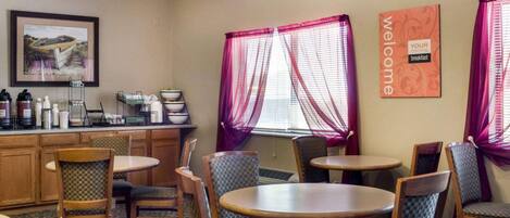 A hotel dining area with wooden chairs and tables, a coffee station, and a 'Welcome' sign.