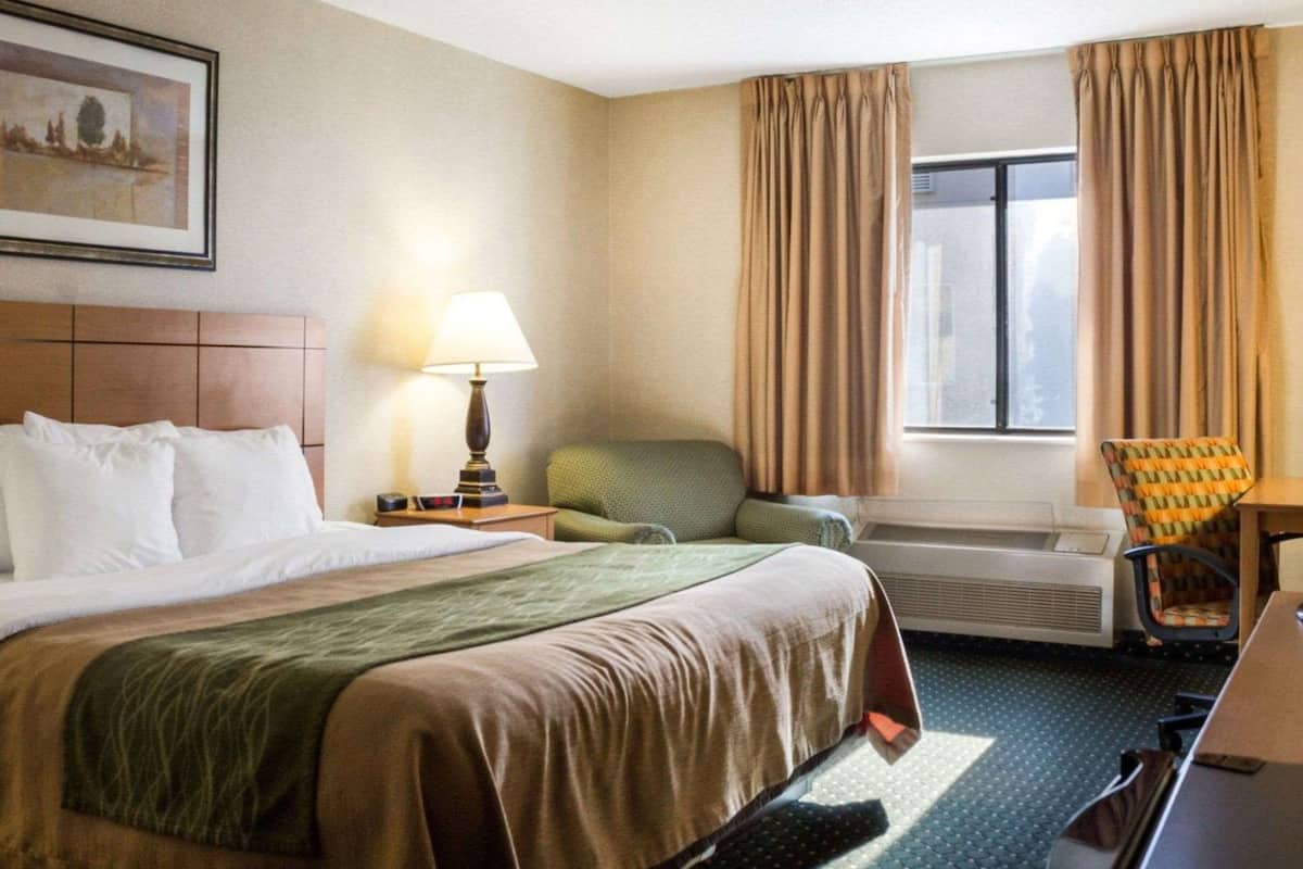 A hotel room with a large bed, a desk, a chair, a window with curtains, and a painting on the wall.