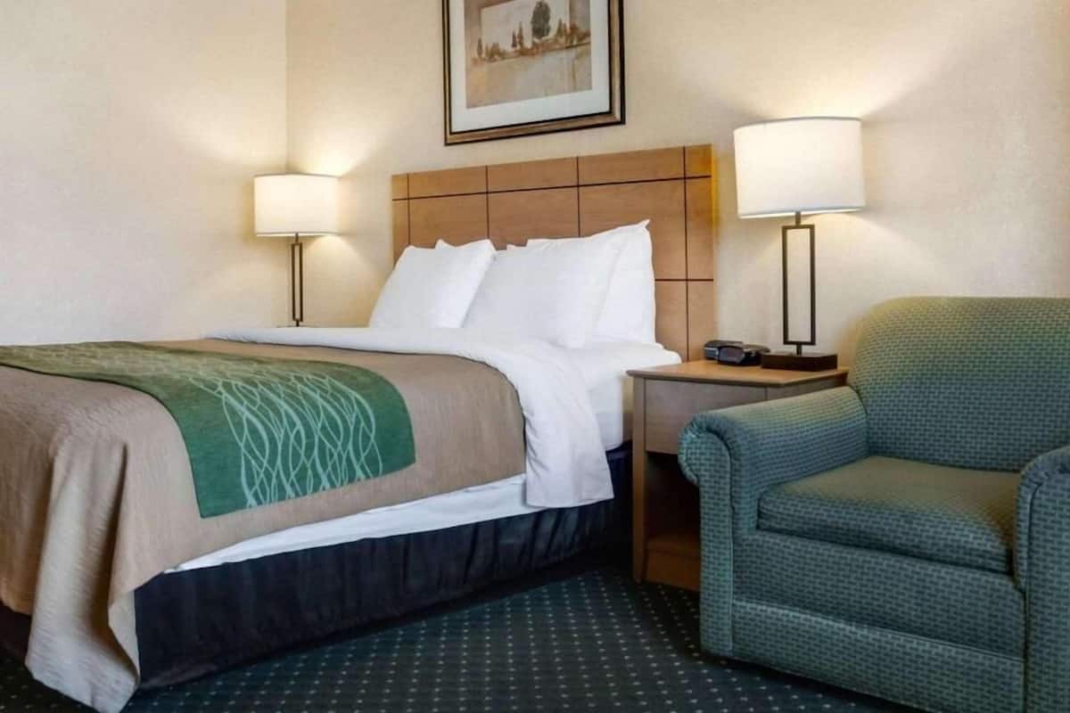 A hotel room with a bed, two bedside lamps, a chair, and a framed picture on the wall.