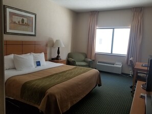 A hotel room with a large bed, a desk, a chair, a lamp, a window with curtains, and a wall-mounted air conditioner. - Comfort Inn Jamestown South (Jamestown)