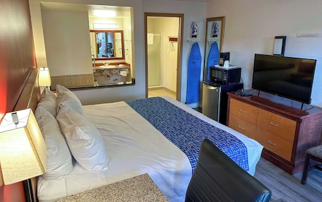 A hotel room with a bed, a desk with a television, a mirror, a shower, and a surfboard.
