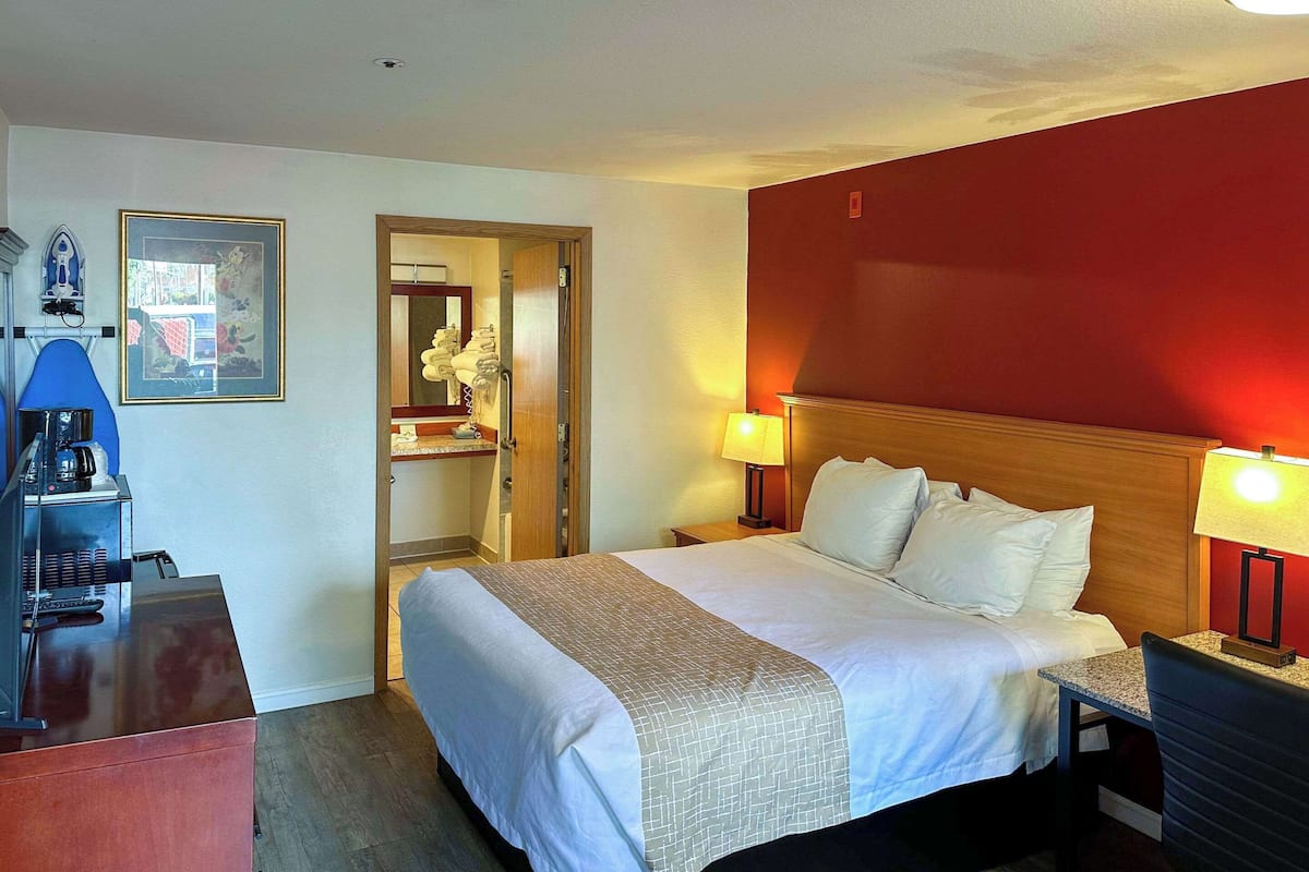 Habitación de hotel con cama, lámparas de noche, escritorio con cafetera, un cuadro enmarcado y espejo.