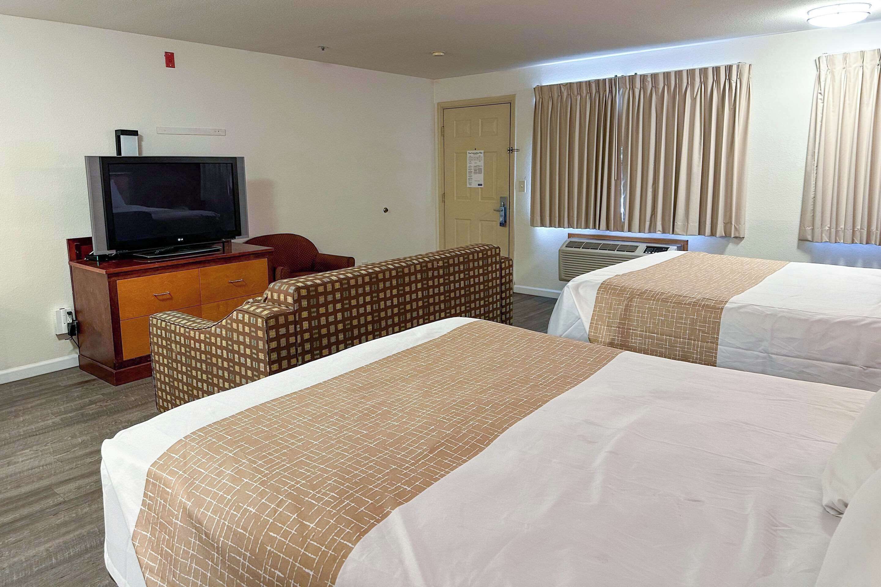 Una habitación de hotel con dos camas, un televisor sobre un aparador de madera, una silla roja y una ventana con cortinas.