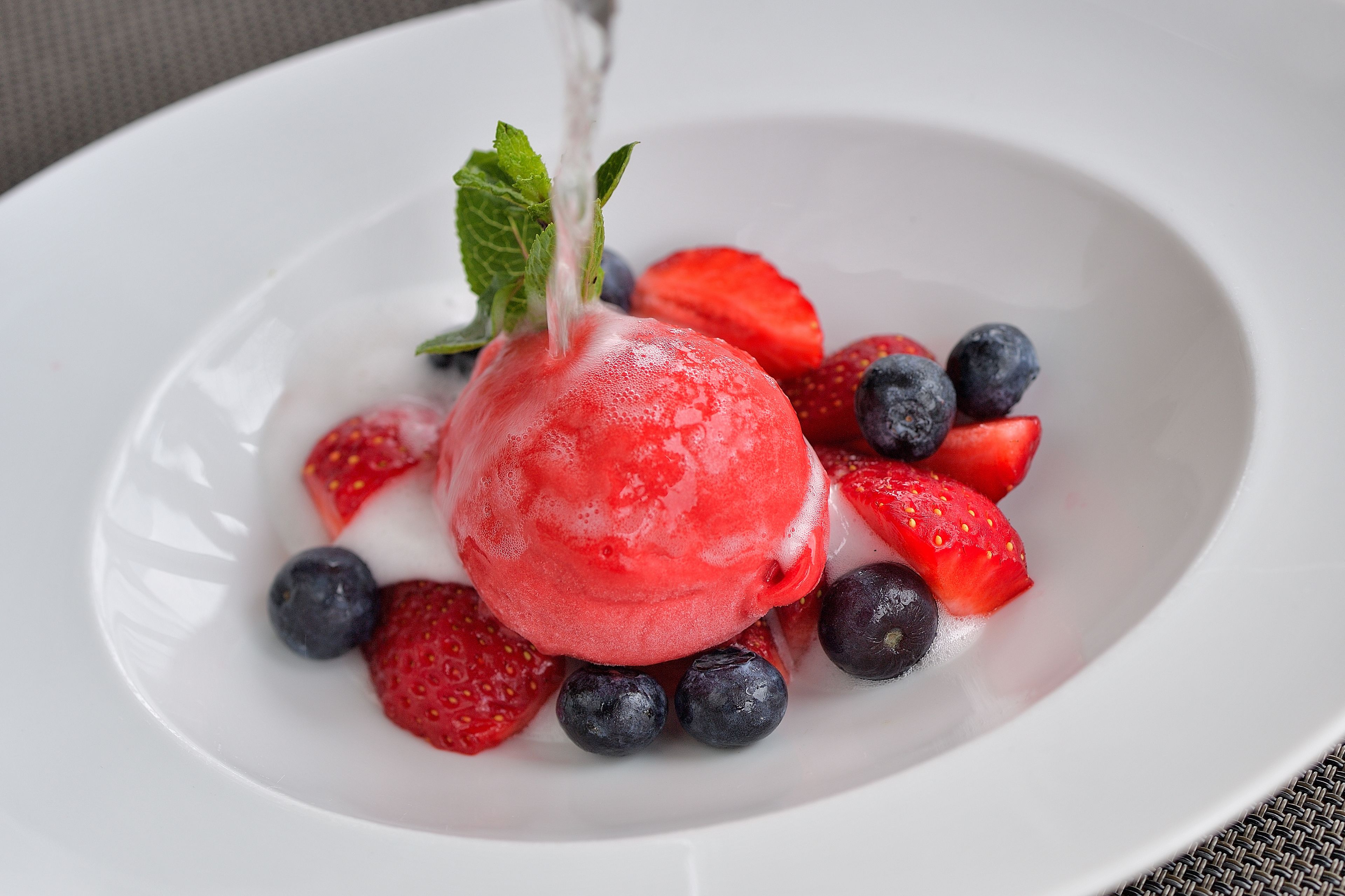 Un postre con una porción de helado rojo rodeada de fresas y arándanos sobre un plato blanco.