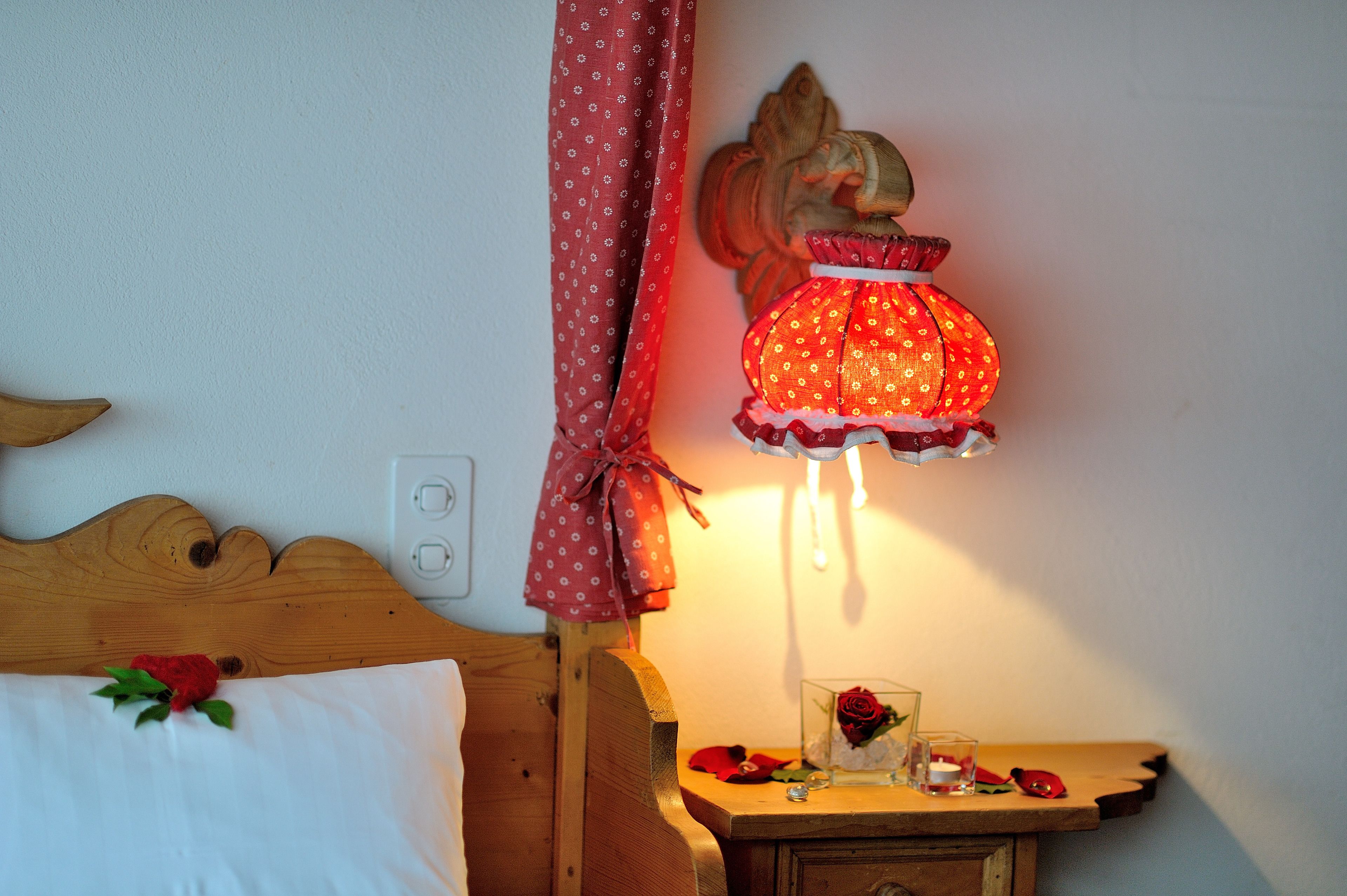 A wooden bed with a white pillow and a red rose. A small wooden table with a glass vase and a red rose is next to the bed.