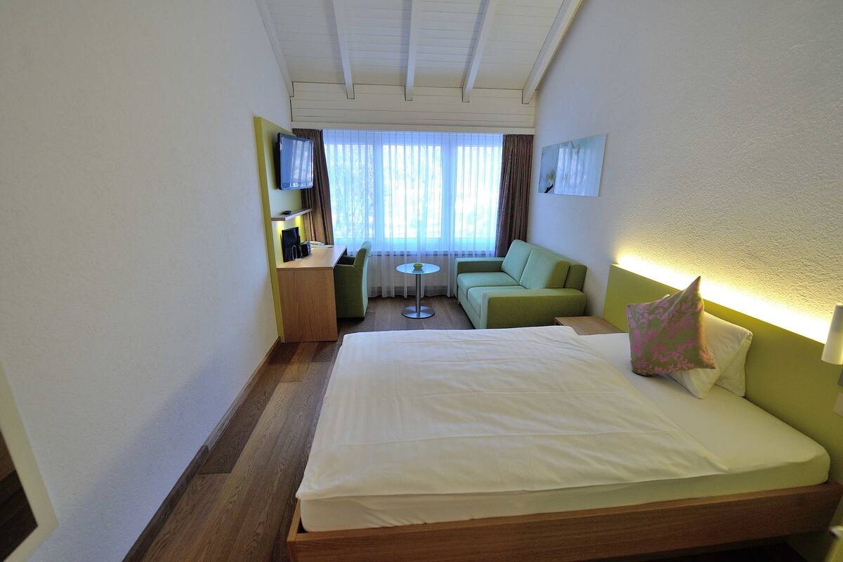 A hotel room with a bed, a green sofa, a small table, a TV, and a window with curtains.