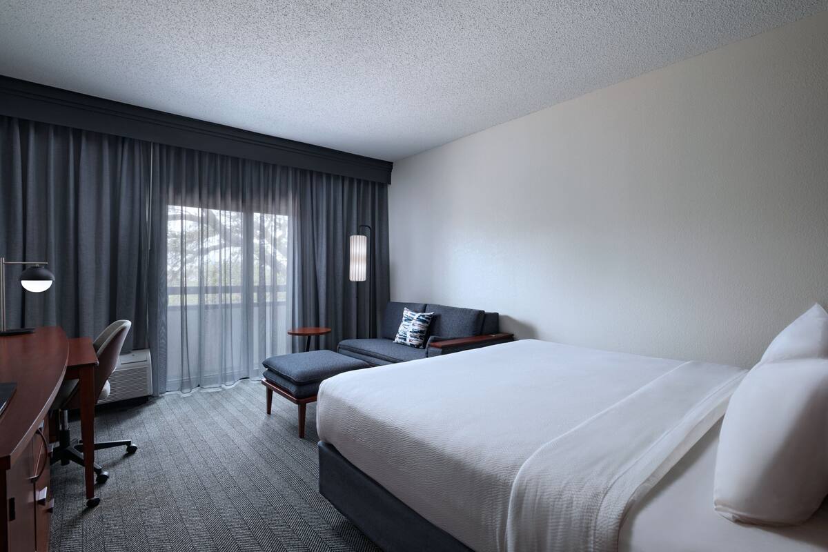 A hotel room with a large bed, a desk, and a chair. There is a window with curtains, a lamp, and a wall-mounted heater.