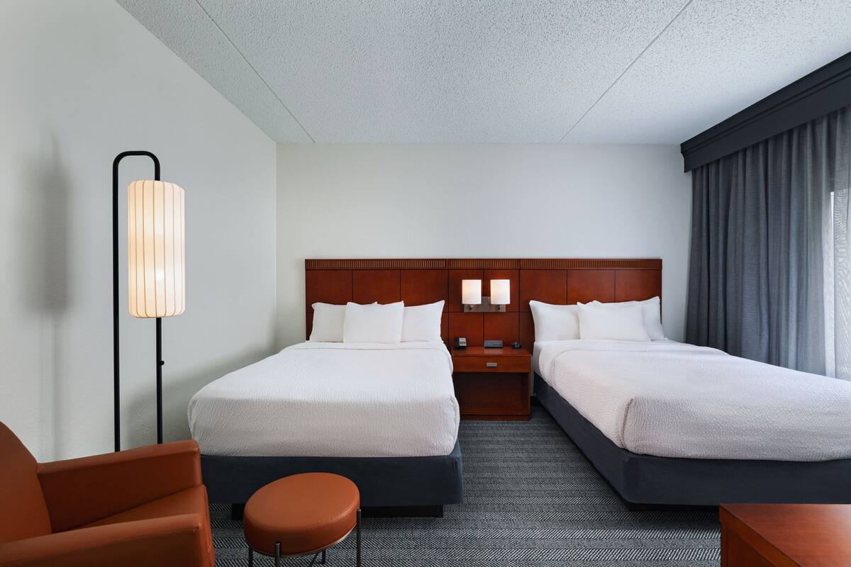 A hotel room with two beds, a desk, a chair, a lamp, and a view of the outside.