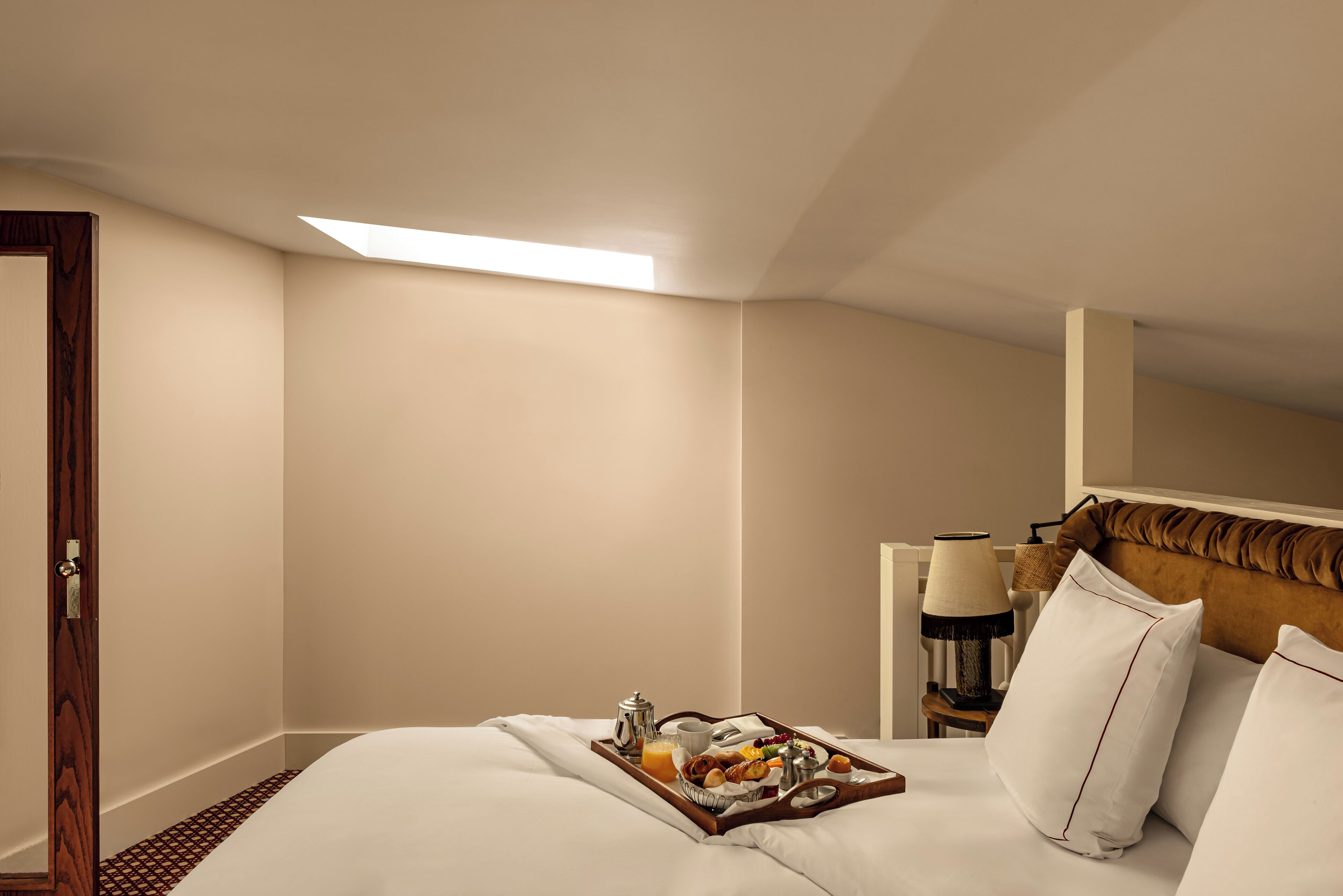 Habitación de hotel con una cama bien tendida, una bandeja con alimentos para el desayuno, una mesita de noche con lámpara y una ventana con luz natural.