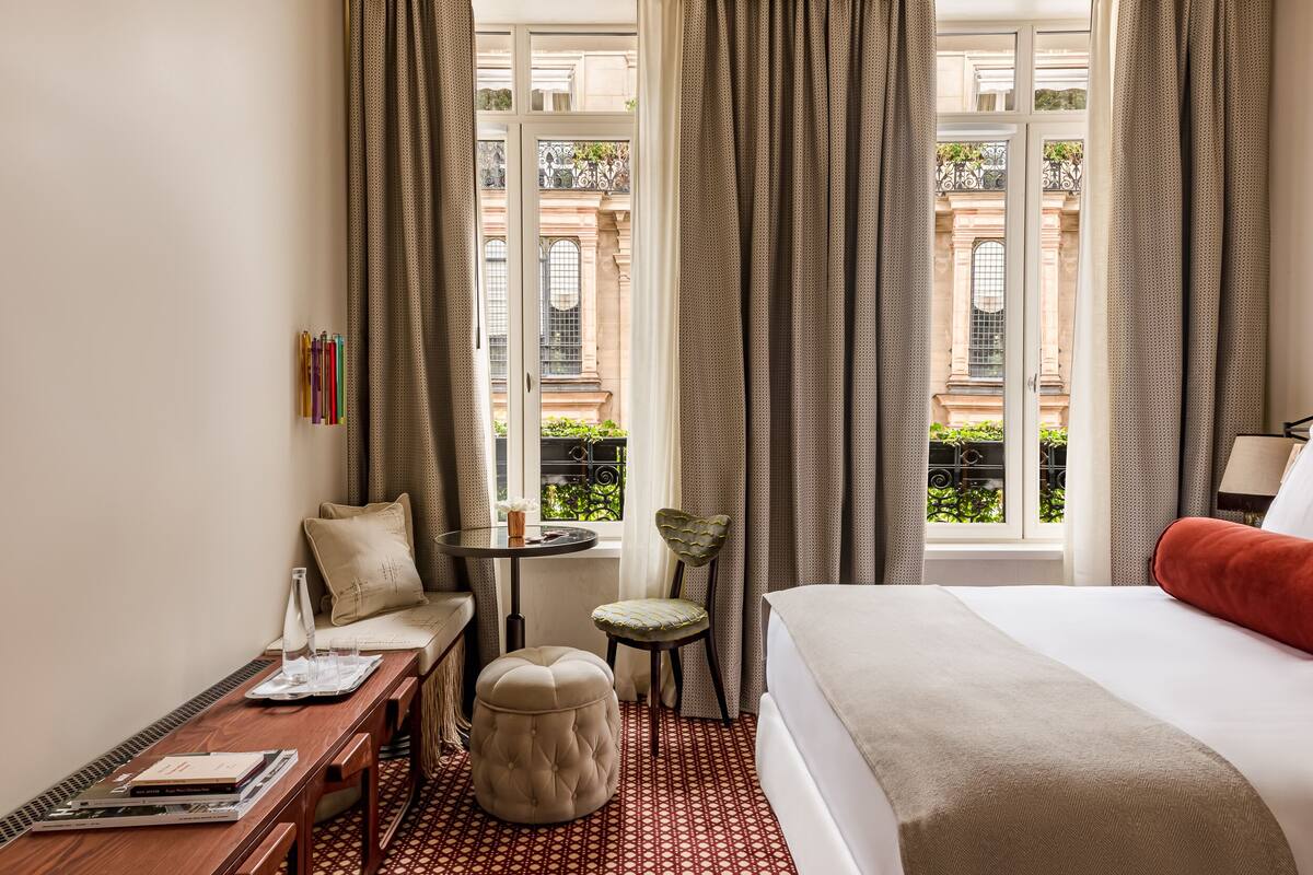 A hotel room with a bed, a small table, a chair, and a window with curtains.