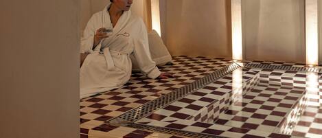 A person in a white bathrobe sitting on a checkered floor in a room with arched ceilings and a tiled wall.