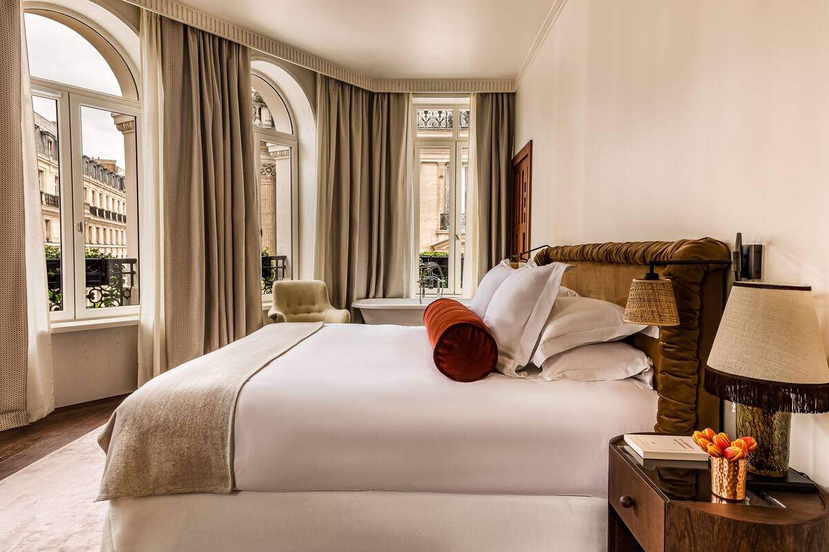 A hotel room with a large bed, a nightstand with a lamp, and a view of the city through large windows.
