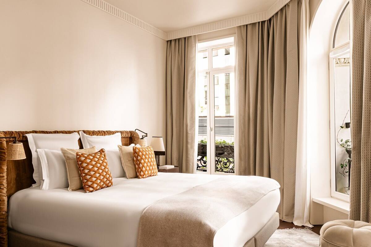 A large bed with multiple pillows, a bedside table with a lamp, and a view of the outside through a window with curtains.