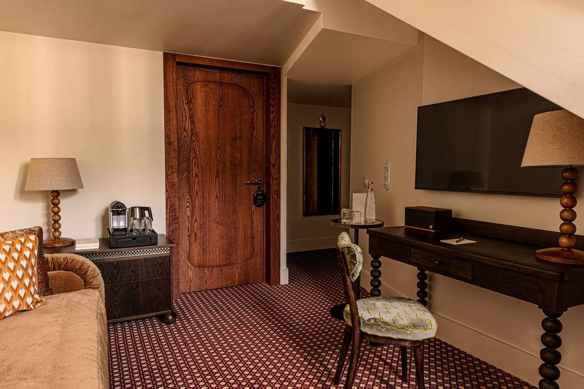 A hotel room with a wooden door, a desk with a lamp, a chair, a bed, and a TV.