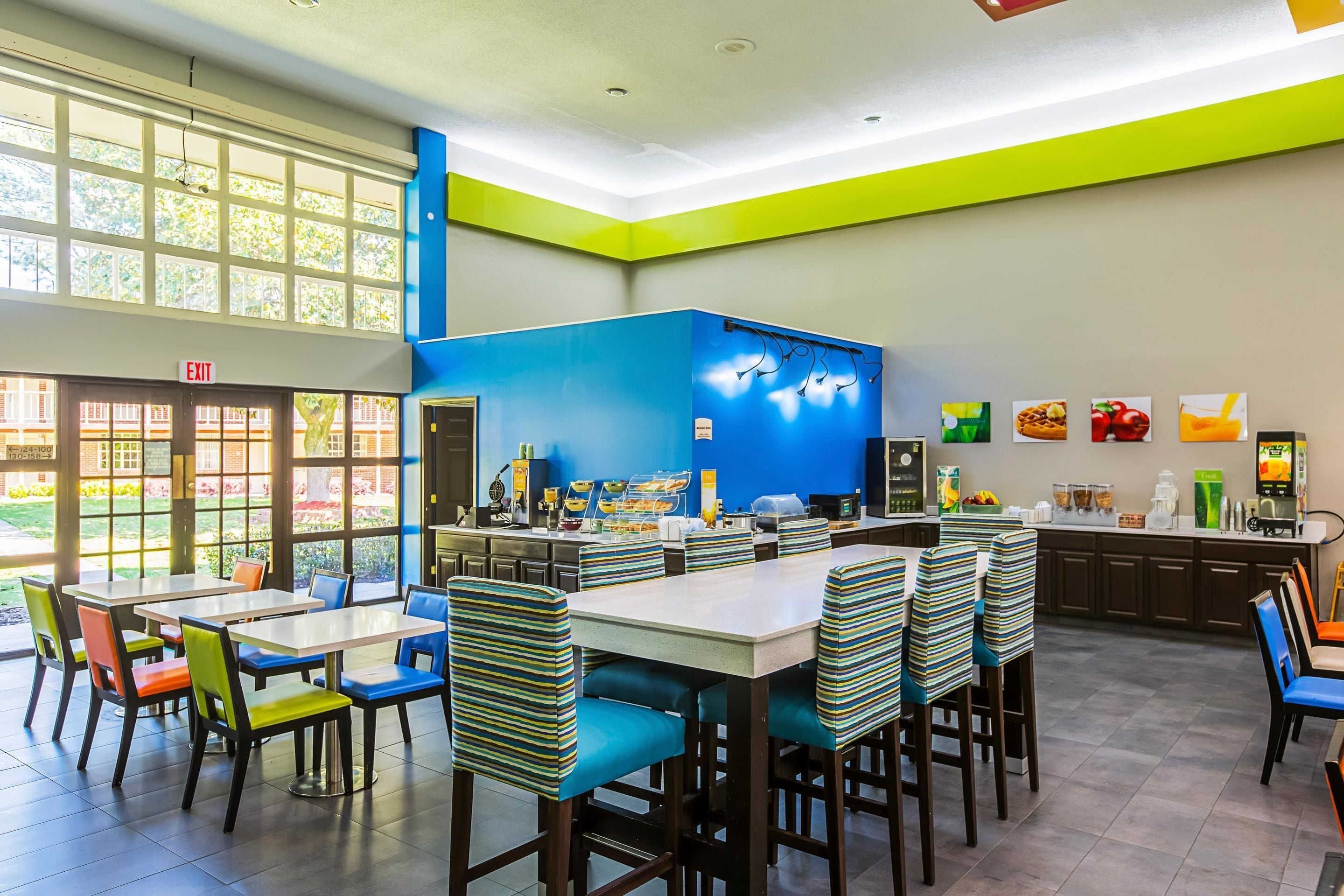 A brightly lit cafeteria with tables and chairs, a kitchen area, and a large window providing natural light.