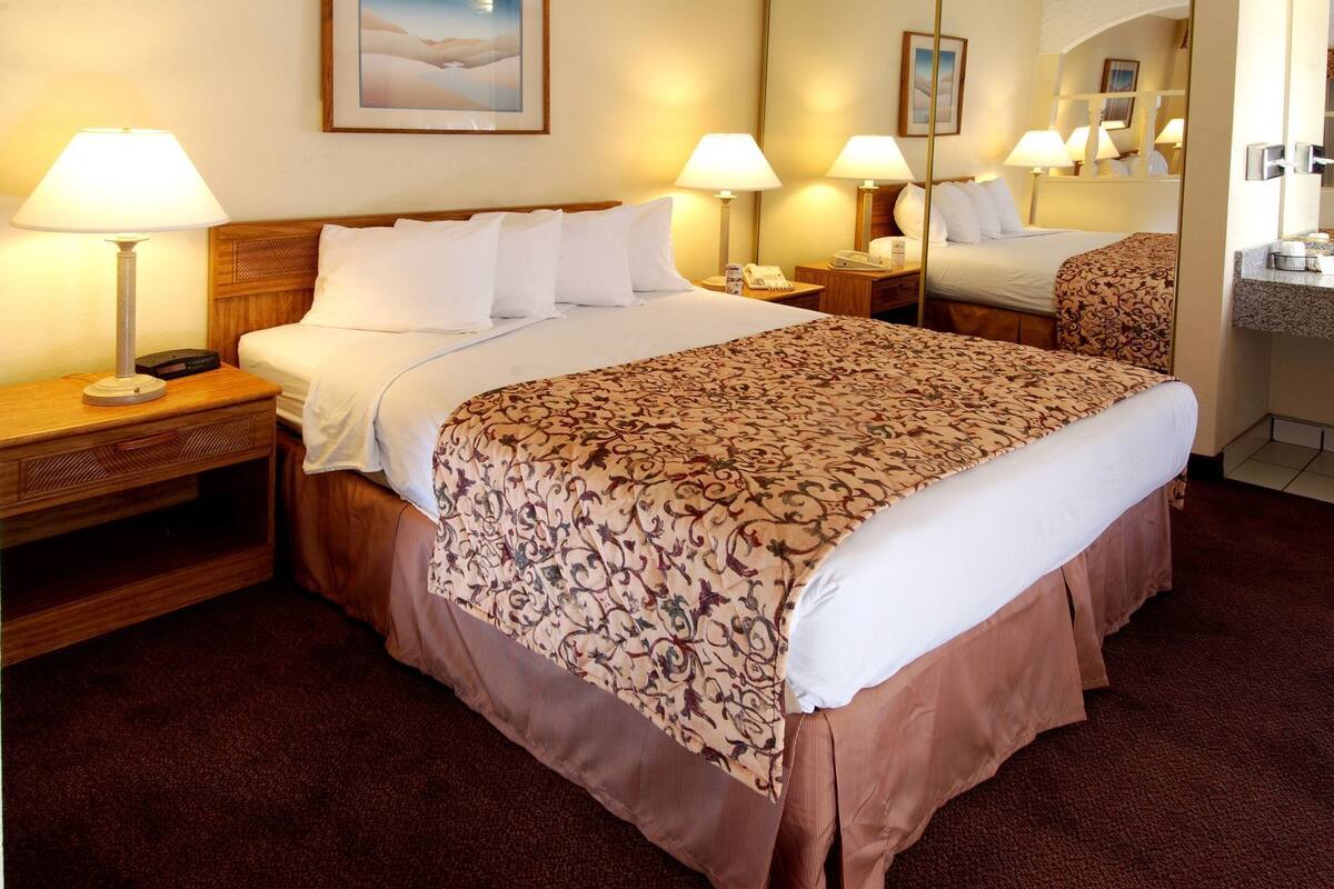 A hotel room with a bed, two bedside tables with lamps, a mirror, a painting on the wall, and a carpeted floor.