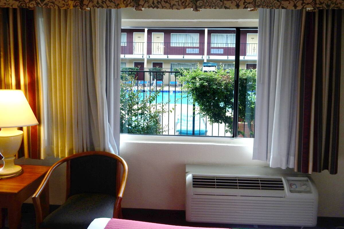 A hotel room with a bed, a chair, a desk with a lamp, an air conditioner, and a window with a view of a building and a pool.