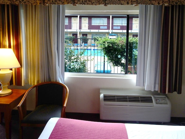 A hotel room with a bed, a chair, a desk with a lamp, an air conditioner, and a window with a view of a building and a pool.