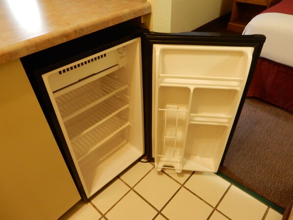 A compact refrigerator with a freezer compartment and a shelf, situated in a small kitchen area with a tiled floor.