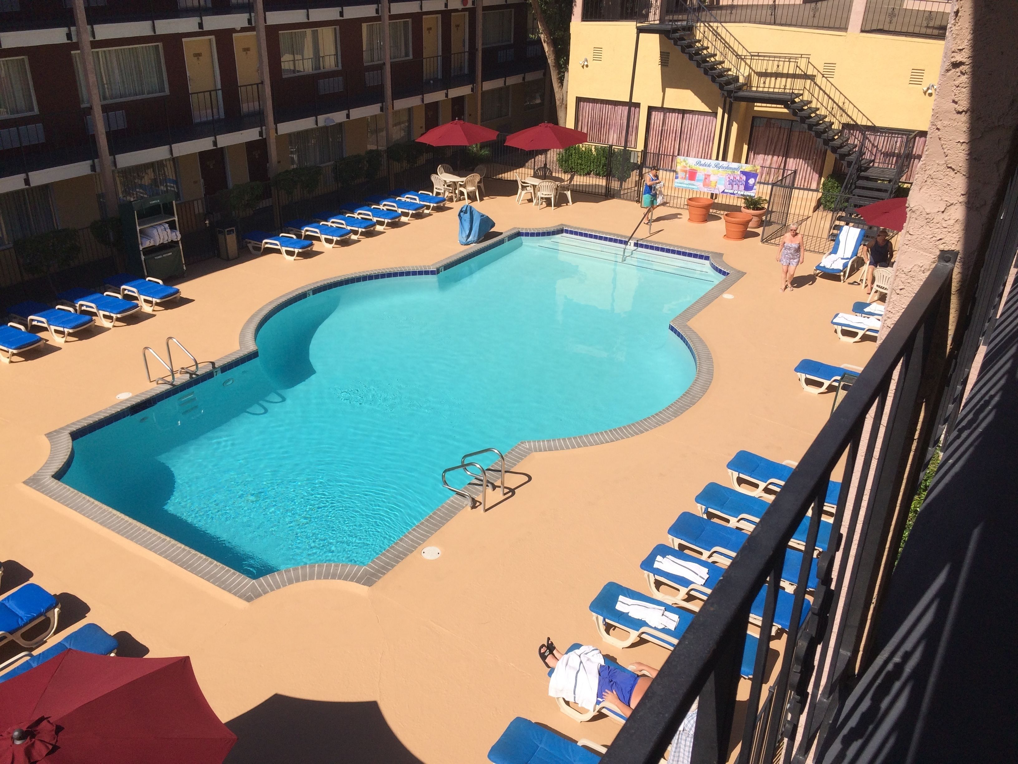 A swimming pool with blue lounge chairs and umbrellas, surrounded by a building with balconies and windows.