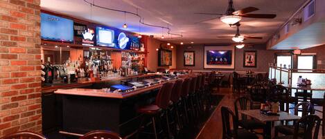 A bar area with a long counter, multiple TVs, and a variety of bottles. There are several tables and chairs arranged for seating.