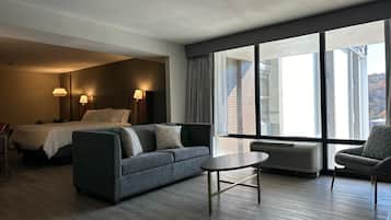 Habitación de hotel con cama, sofá, una mesita y una silla. Se aprecia un ventanal con vistas a edificios y un muro de ladrillo.