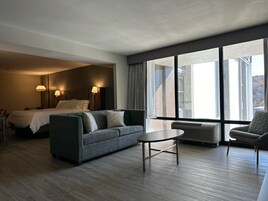Habitación de hotel con cama, sofá, una mesita y una silla. Se aprecia un ventanal con vistas a edificios y un muro de ladrillo.