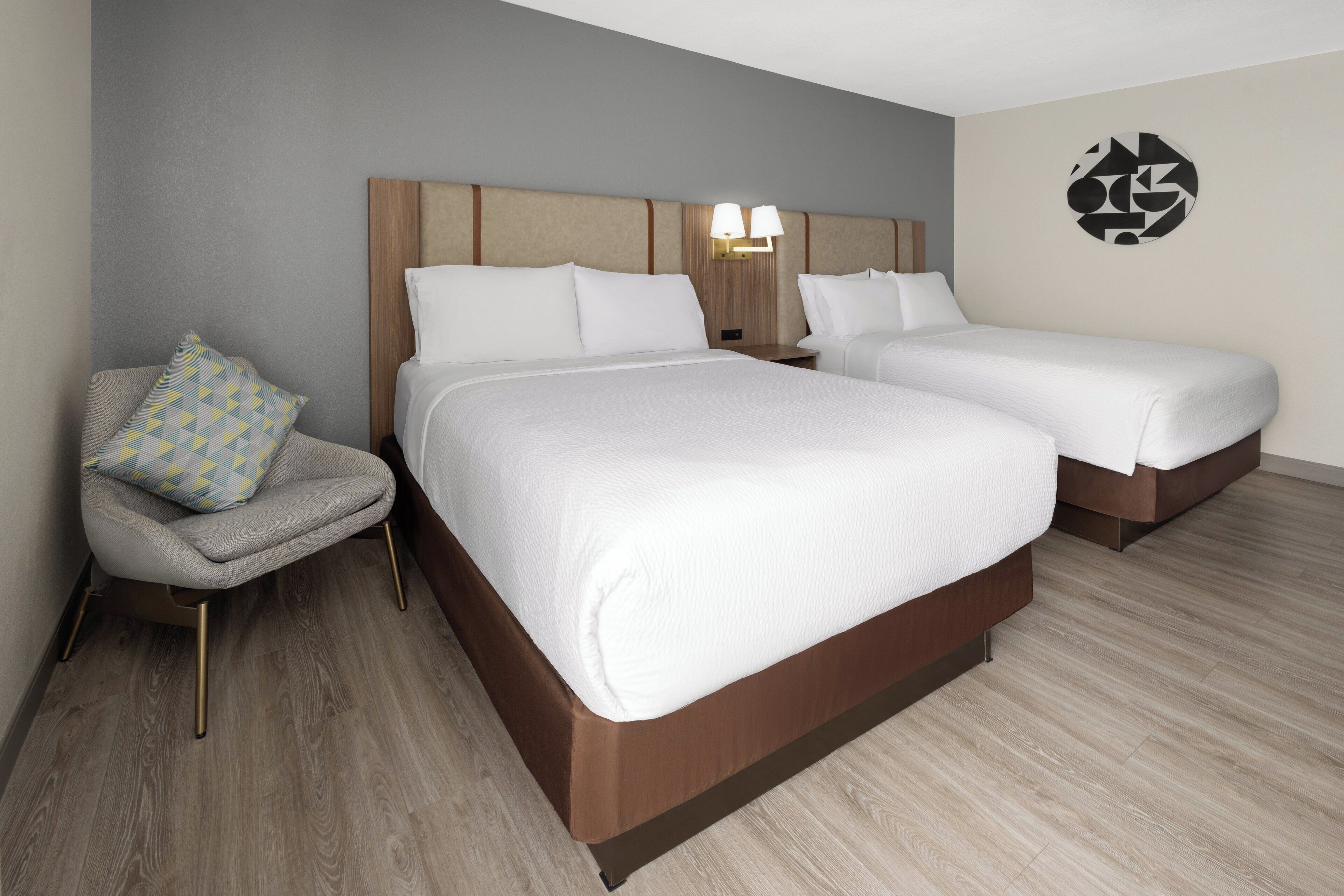 A hotel room with two beds, a nightstand with lamps, a chair with a patterned cushion, and a wall art piece.