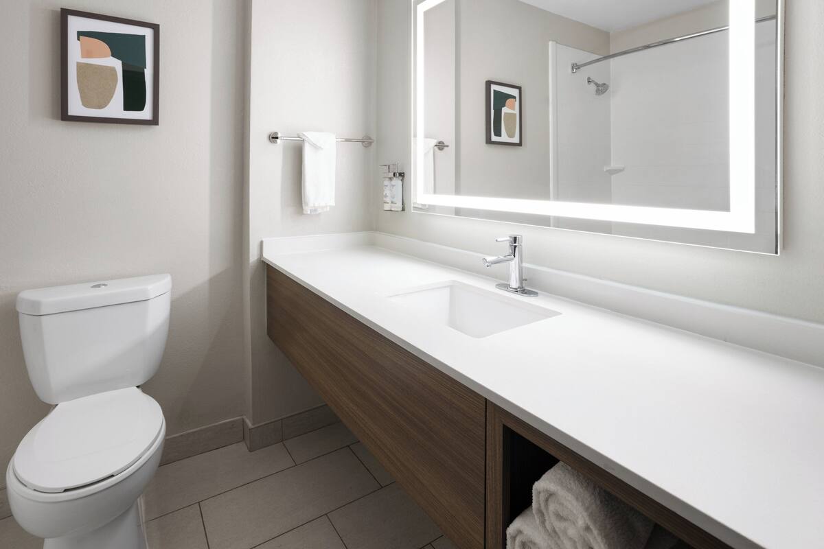 Une salle de bain moderne avec des toilettes blanches, un meuble-lavabo en bois, un grand miroir et une œuvre d’art encadrée accrochée au mur.