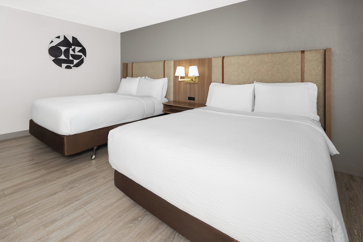 A hotel room with two beds, a wooden headboard, a wall-mounted light fixture, and a circular wall art piece.