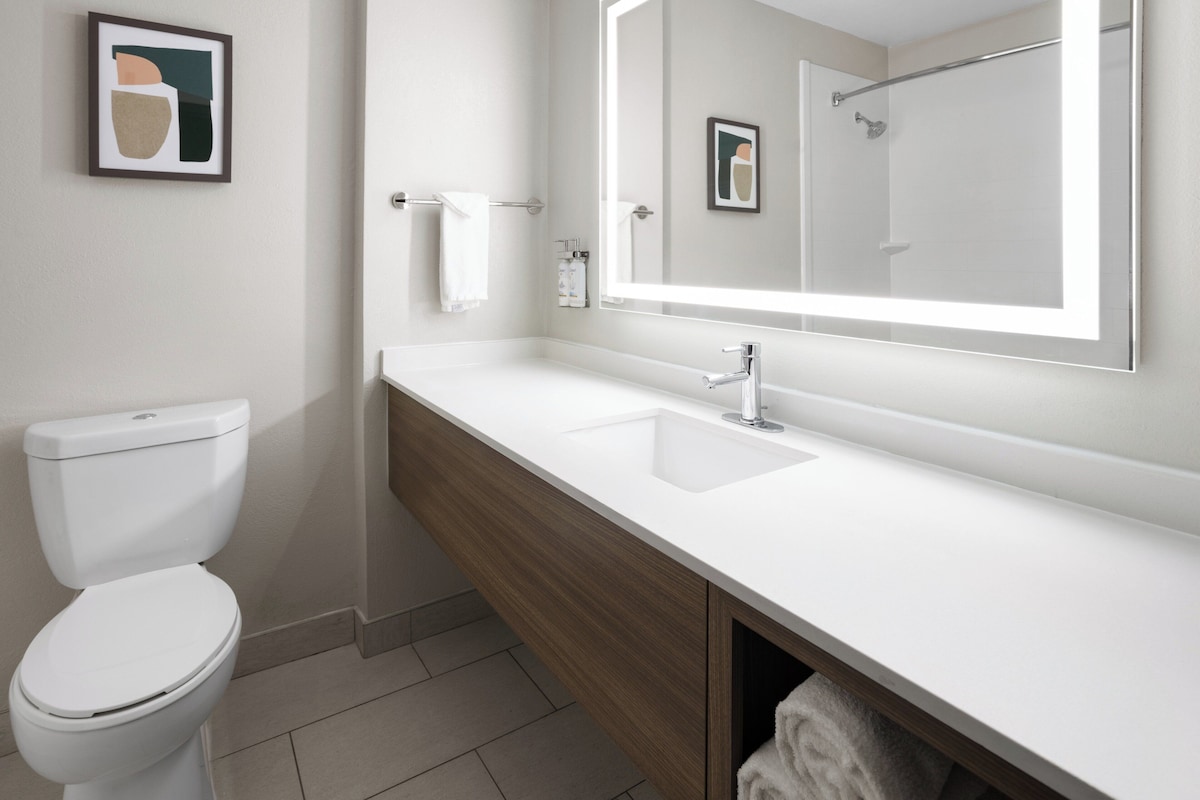 A modern bathroom with a white toilet, a wooden vanity, a large mirror, and a framed artwork on the wall.