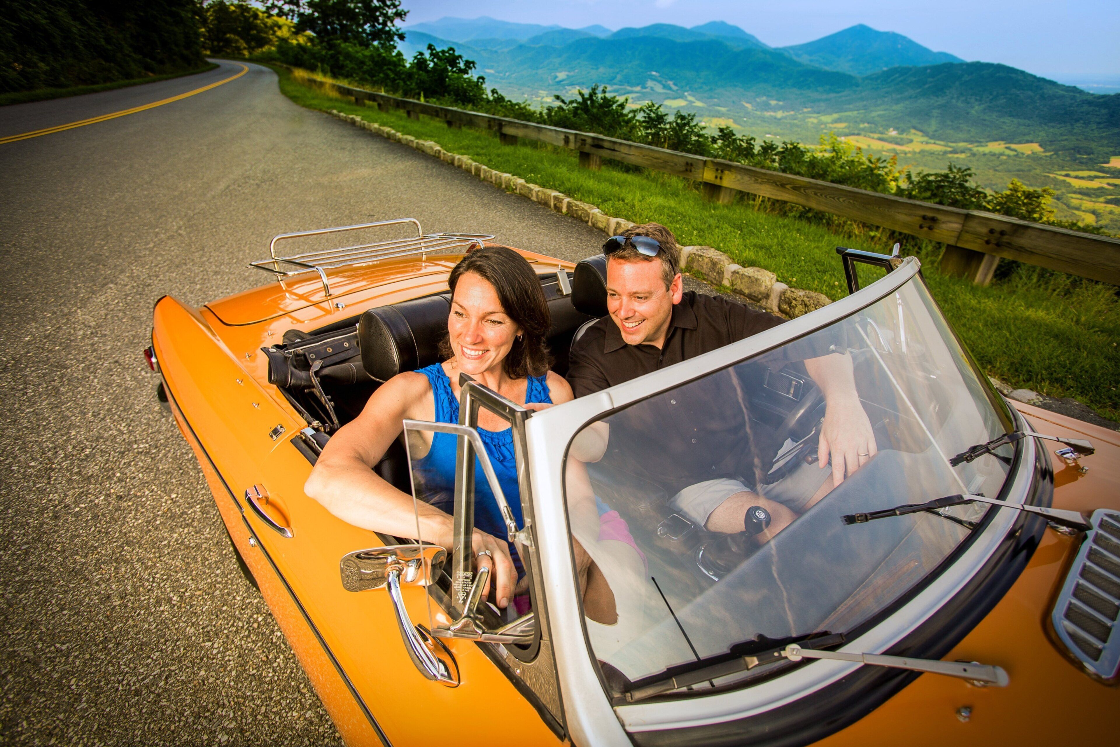 A convertible car on a road with a scenic view of mountains and greenery.