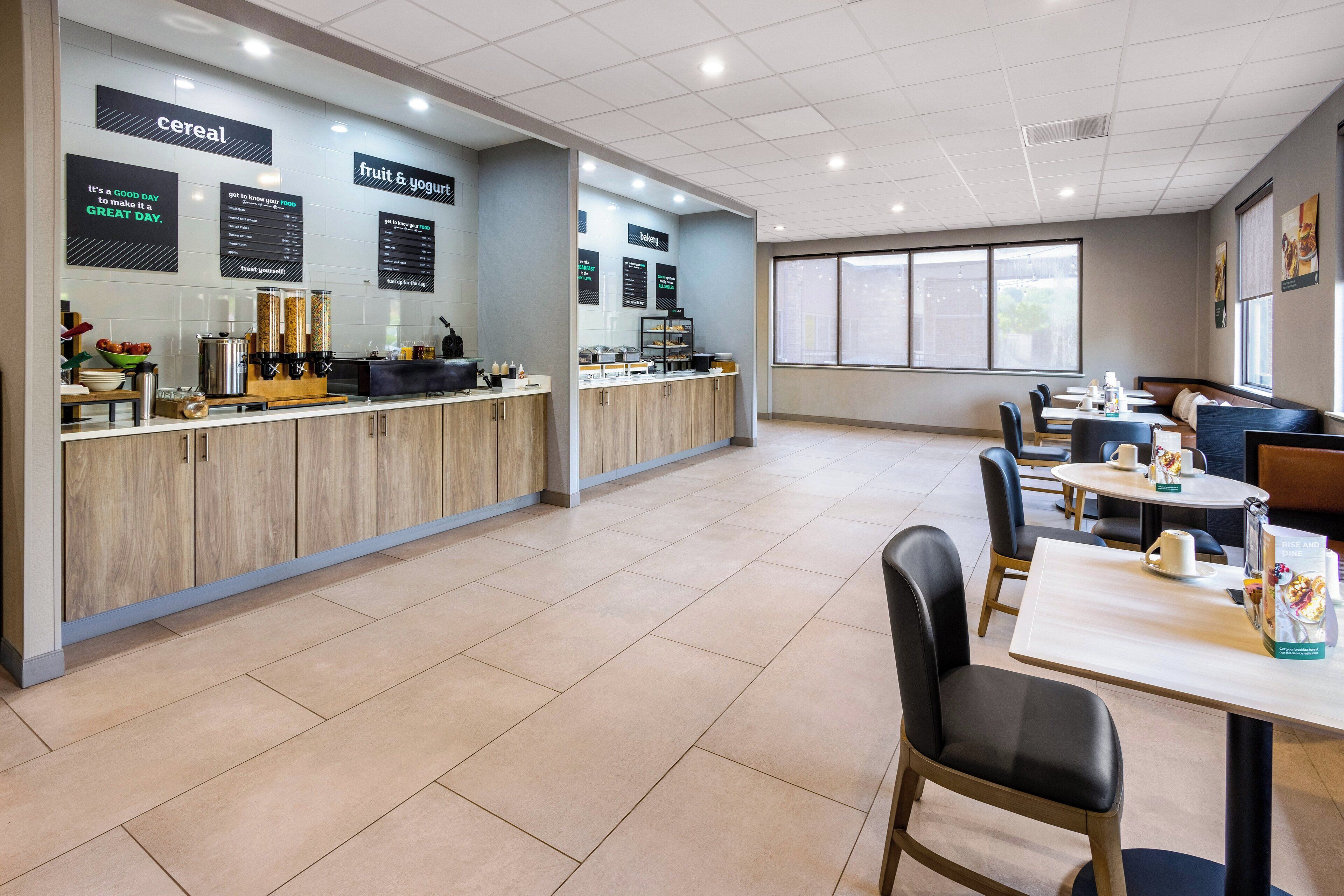Un café moderne avec un comptoir où sont exposés les articles proposés pour le petit-déjeuner, un espace salon avec des tables et des chaises, et une grande fenêtre qui laisse entrer la lumière naturelle.